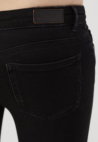 Vaqueros de mezclilla negra con una vista en primer plano del bolsillo trasero. Presenta una etiqueta de marca de cuero marrón y detalles de costura sutiles.