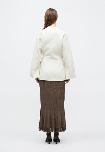 Λευκό σακάκι με προσαρμοσμένη ραφή, καμπυλωτά μανίκια και εφαρμοστή μέση, συνδυασμένο με μακρύ καφέ φούστα με βολάν, επίπεδα και δαντέλα.