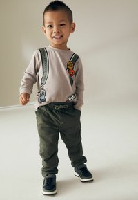 Chemise à manches longues gris clair avec des bretelles imprimées et des patchs ; pantalon à taille élastique vert olive ; baskets noir et blanc.