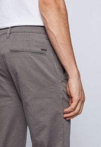Grå chinos med en slät textur, med en bakficka, diskreta sömmar och en liten logotypetikett. Enkel design utan synliga mönster.