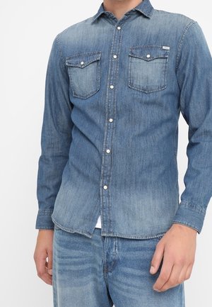 Chemise en denim dans des tons de bleu variés, avec un col, un boutonnage sur le devant et deux poches poitrine. Coupe ajustée avec des manches longues.