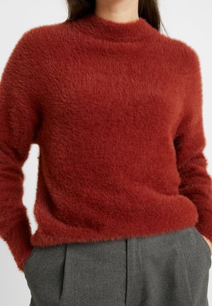 Pull rouge en laine à col montant et manches longues, avec une texture douce. Associé à un pantalon gris ajusté.