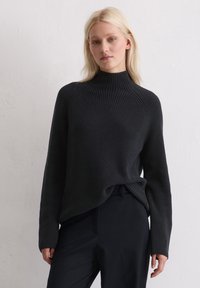 Pull à col roulé noir à côtes, avec des manches longues, présentant un design tricoté texturé et une coupe décontractée, associé à un pantalon sombre.
