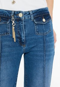 Vaqueros de mezclilla azul con una cintura de altura media, bolsillos frontales, acentos de hardware en dorado y un charm de borla en el tirador de la cremallera. Textura suave.