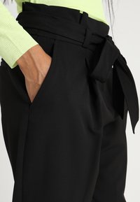 Mustat räätälöidyt housut, joissa on leveä vyötärö, solmittava vyö ja sivutaskut. Kangas näyttää sileältä ja istuu muotoon.