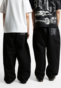 Black baggy jeans with prominent back pockets featuring embroidered designs, paired with a white and a graphic black shirt.