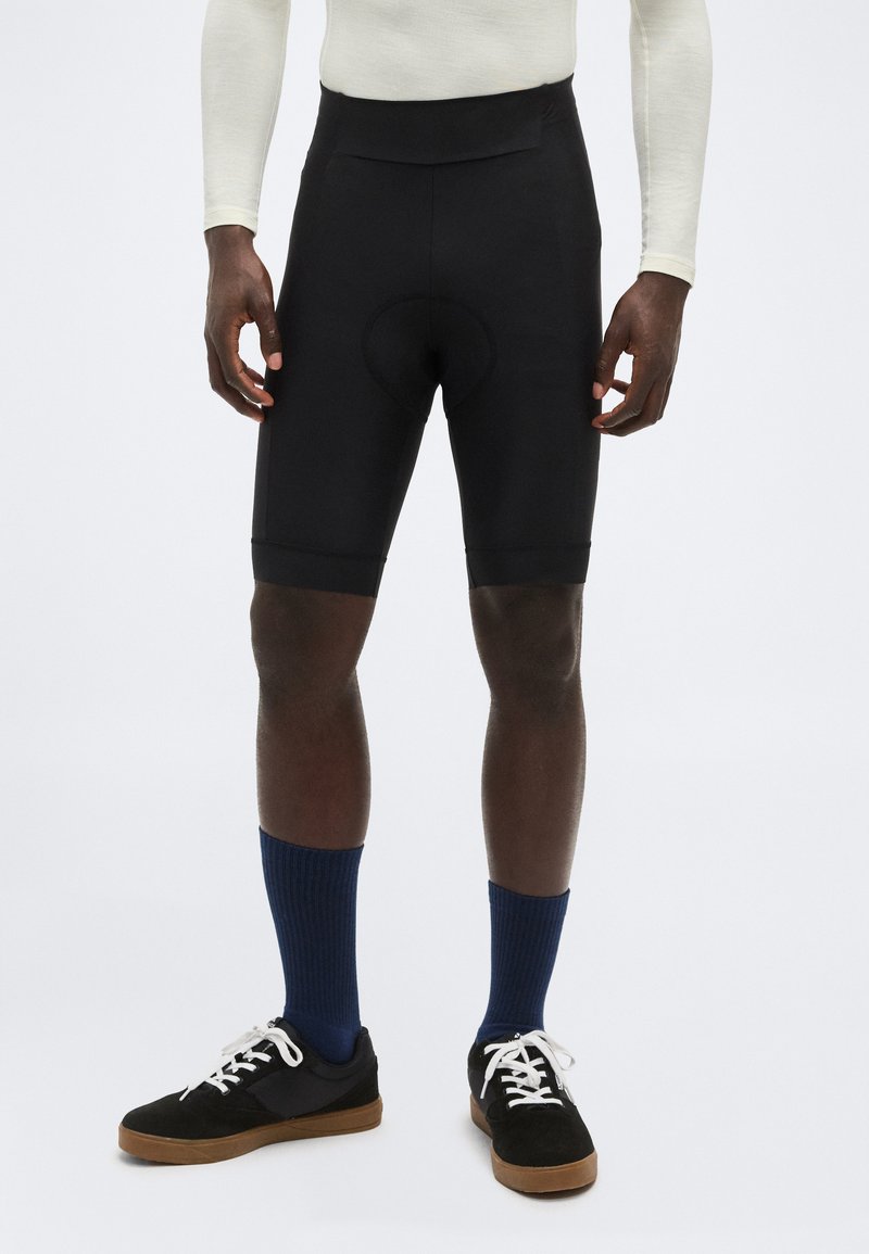 Un homme porte un cuissard rembourré noir, des chaussettes bleu marine, des baskets noires et un haut à manches longues crème, debout devant un fond blanc.