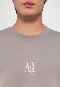 T-shirt in cotone grigio con collo a giro, con un logo ricamato bianco per "Armani Exchange" nella parte sinistra del petto.