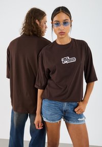 Camiseta oversize marrón con un estampado gráfico blanco, combinada con pantalones cortos de denim. La tela parece suave y tiene un corte relajado.