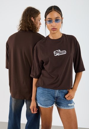 Rjav pobijan t-shirt z belim grafičnim tiskom, v kombinaciji z denim kratkimi hlačami. Tkanina se zdi mehka in sproščena.