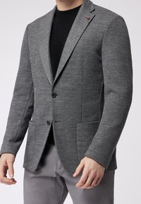 Uomo che indossa un blazer grigio a coste con bottoni neri, un maglione nero a girocollo e pantaloni grigi, in piedi di fronte a uno sfondo uniforme.
