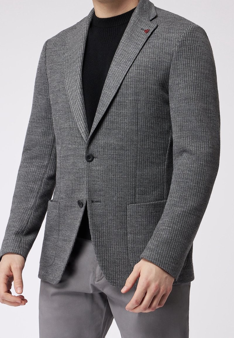 Uomo che indossa un blazer grigio a coste con bottoni neri, un maglione nero a girocollo e pantaloni grigi, in piedi di fronte a uno sfondo uniforme.