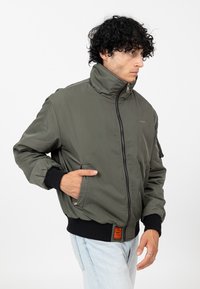 Veste bombardier vert olive avec col montant, fermeture éclair et poignets côtelés noirs. Dispose de poches latérales et d'un détail en écusson sur la manche.