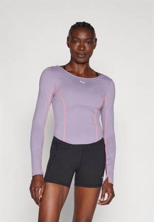 Camiseta deportiva de manga larga en lila claro con detalles naranjas, con un diseño ajustado. Combinada con shorts negros, que muestran un material atlético.