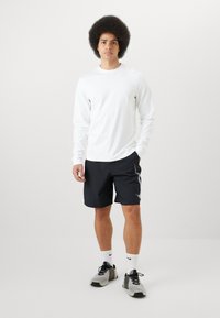 Chemise blanche à manches longues, shorts de sport noirs avec des rayures blanches sur le côté, chaussures de sport grises et chaussettes blanches. Le modèle se tient contre un fond uni.