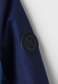 Tela texturizada de color azul marino con un parche redondo de cuero oscuro que presenta un logo y detalles en relieve. Diseñado para la durabilidad y el estilo.