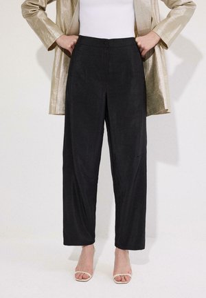 Kobieta ubrana w czarne, szerokie spodnie z wysokim stanem, białą bluzkę, złotą marynarkę i białe sandały na obcasie z odkrytymi palcami, stojąca z rękami na biodrach.