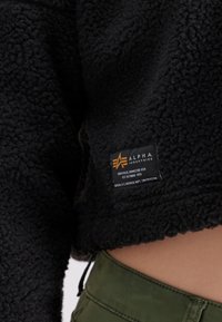 Svart fleecegenser med en teksturert, myk overflate. Har et merke med "Alpha Industries"-logo og pleiedetaljer på nedre kant.