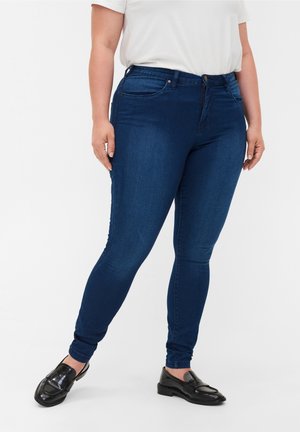 Person iført mørkeblå skinny jeans med forlommer og sorte slip-on loafers, stående mod en ensfarvet hvid baggrund.