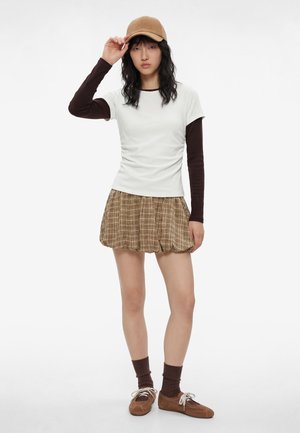 Jeune femme portant une casquette beige, une chemise blanche à manches courtes sur un sous-pull marron à manches longues, une jupe à carreaux marron, des chaussettes marron et des chaussures marron, debout.
