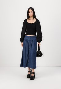 Blusa nera a maniche lunghe con texture arricciata, abbinata a una gonna in denim blu con bottoni. Il modello tiene una piccola borsa nera e indossa scarpe chunky con plateau.