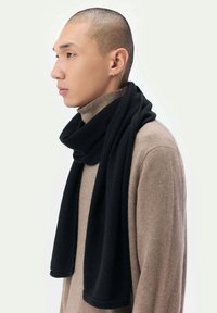 Svart scarf som bärs draperad runt halsen över en ljusbrun tröja, med ett mjukt, strukturerat tyg och en enkel, elegant design.