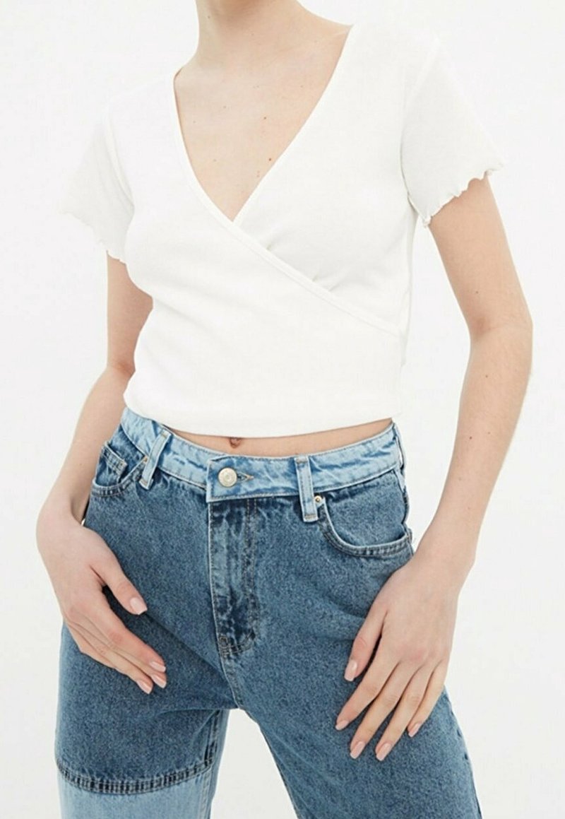 Top avvolgente bianco corto con maniche corte e bordi ondulati, abbinato a jeans blu a vita alta con un design in denim chiaro e scuro.