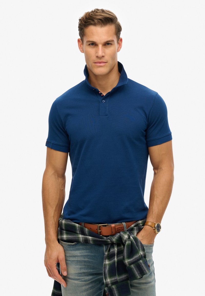 Polo shirt blu navy con colletto corto e due bottoni, con polsini arrotolati. Indossato con una camicia a quadri legata in vita.