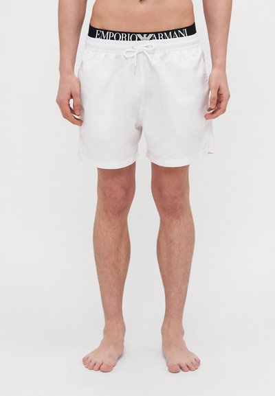 Shorts de bain blancs avec une taille élastique et un cordon de serrage noir ; présente une bande de logo noire. Tissu léger, longueur au-dessus du genou, design sobre.