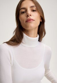 Weißer gerippter Rollkragenpullover mit einem halbtransparenten Design und einer taillierten Silhouette. Glatte Textur mit hohem Kragen und langen Ärmeln.
