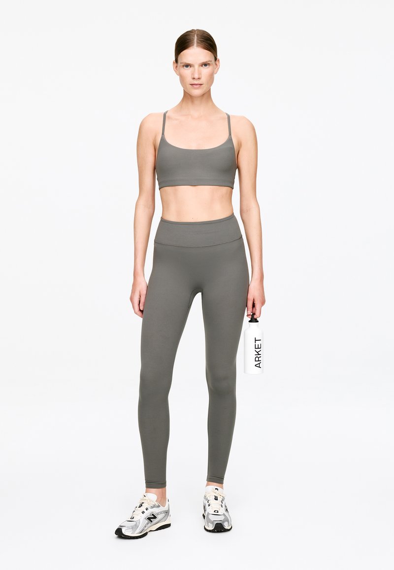 Femme en brassière de sport grise et leggings tenant une bouteille d'eau blanche avec le texte « ARKET », portant des chaussures de course blanches et grises, debout sur un fond blanc.
