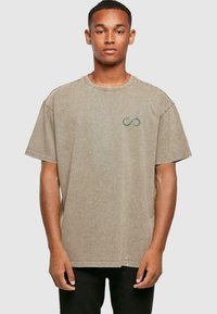 Beige bomulls t-skjorte med rund hals og løst snitt. Har subtil grønn uendelighetssymbol broderi på brystet. Korte ermer.
