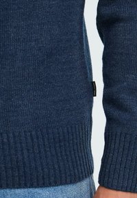 Tengerészkék kötött pulóver bordás mandzsettával és aljjal. Oldalán található egy kis márkajelzés, amely puha textúrát és testhezálló fazont mutat.