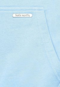 Light blue hoodie fabric with a soft texture, featuring a front pocket and a silver logo tag on the side. Simple design and stitching details.