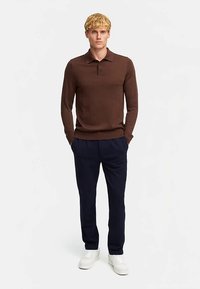 Polo marrone a maniche lunghe con colletto, abbinata a pantaloni blu scuro. Il modello indossa sneakers bianche, in piedi davanti a uno sfondo semplice.