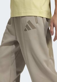 Beige urheilualueet teksturoidulla pinnalla, joissa on sivutasku ja sävyttävä Adidas-logo yläreidessä. Kapeasti muotoiltu malli.