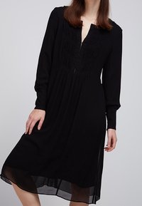 Czarna sukienka o długości średniej z długimi rękawami, teksturowanym bodicem i krótkim zapięciem na guziki. Miękki materiał o zwiewnym kroju.
