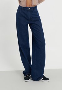 Vaqueros de pierna ancha en tela de mezclilla azul oscuro, con un diseño clásico de cinco bolsillos y una cinturilla de altura media. Combinados con zapatillas negras.