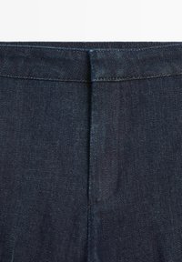 Jean en denim bleu foncé en gros plan montrant la ceinture et la couture centrale avant sans bouton ni fermeture éclair visible.
