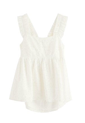 Pige iført hvid ærmeløs top med flæser og matchende bukser med hulmønster, stående med hånden på hoften foran en ensfarvet baggrund.