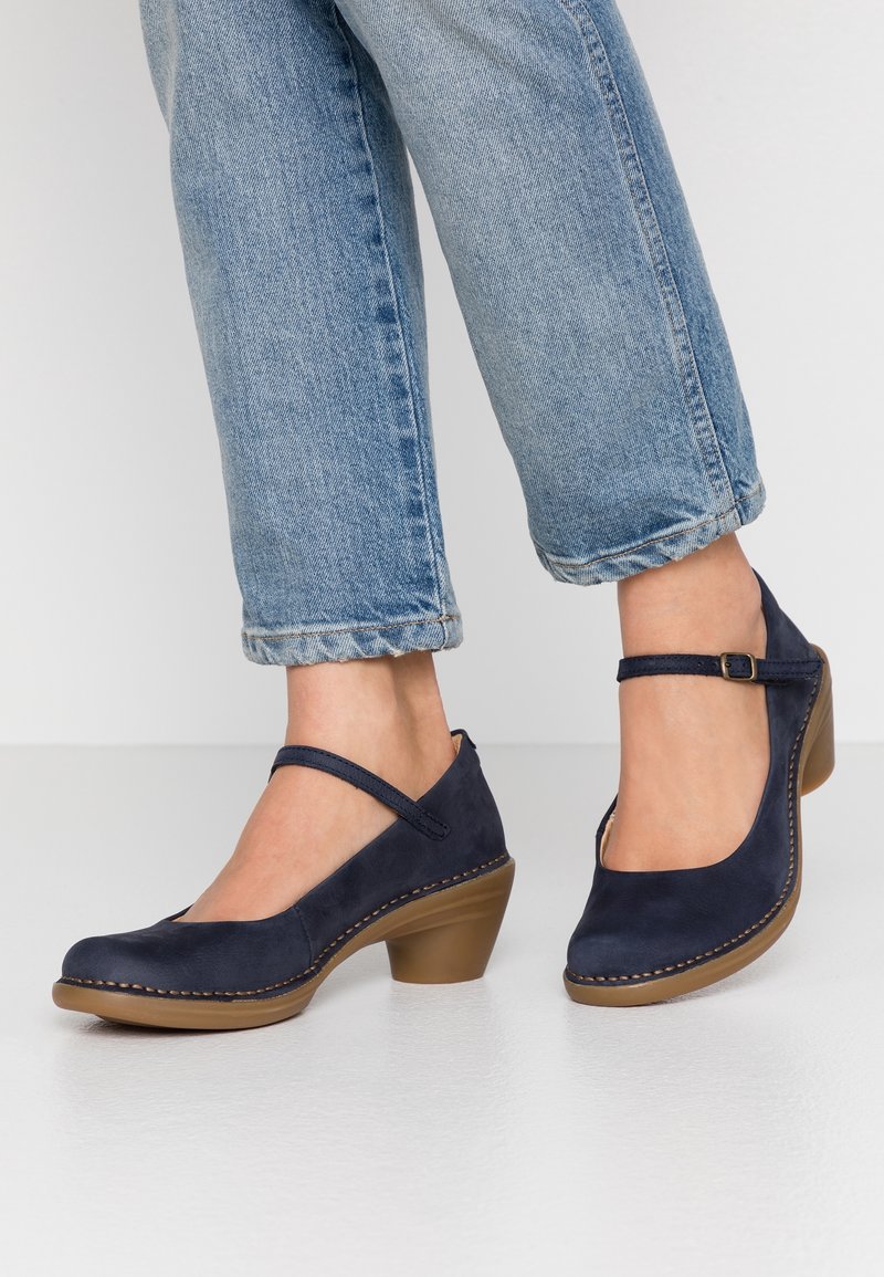 Pieds portant des escarpins Mary Jane en daim bleu marine avec talons blocs marron clair et brides à la cheville, associés à un jean bleu clair retroussé.
