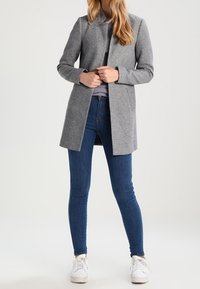 Abrigo gris de mezcla de lana con un diseño sin cuello, bolsillos laterales, combinado con jeans ajustados azul oscuro y zapatillas blancas.