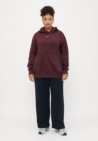 Sweatshirt cor burguês com cordões e bolso canguru, combinado com calças largas azuis escuras, usadas com sapatilhas de atletismo cinza claro.