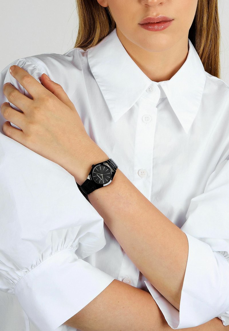 Orologio nero con quadrante rotondo elegante e dettagli argentati, con cinturino nero. Indossato su un polso con una camicia bianca abbottonata.