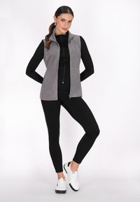 Gilet in pile grigio senza maniche con colletto alto, chiusura zip e texture liscia; indossato sopra una maglietta nera a maniche lunghe e leggings.