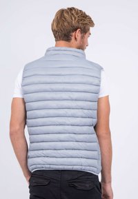 Gilet rembourré gris avec un motif de couture horizontal, un col haut et un matériau léger. Pas de matériel visible. Porté par-dessus une chemise blanche.