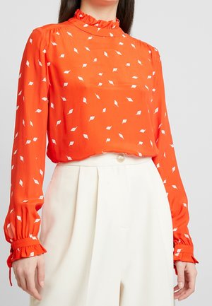 Femme portant un chemisier orange à manches longues avec des motifs de feuilles blanches et des poignets à volants, associé à un pantalon blanc taille haute avec un bouton.