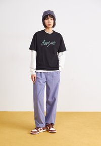 Camiseta negra de algodón con el texto verde "Carhartt", superpuesta sobre una camiseta de manga larga blanca. Combinada con pantalones morados claros y zapatillas marrones.