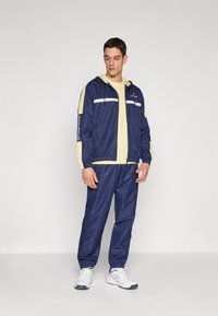 Marine blå joggedrakt med lysegule detaljer, inneholdende en hettejakke med glidelås og elastiske mansjetter. Hvite striper på brystet. Brukt med hvite sportssko.