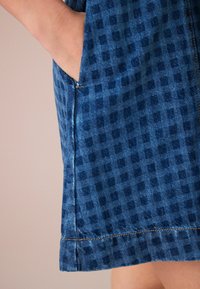 Denim obleka s modrim karirastim vzorcem, sprednjimi žepi in kontrastnimi šivi na robu. Tekstura se zdi rahlo strukturirana.